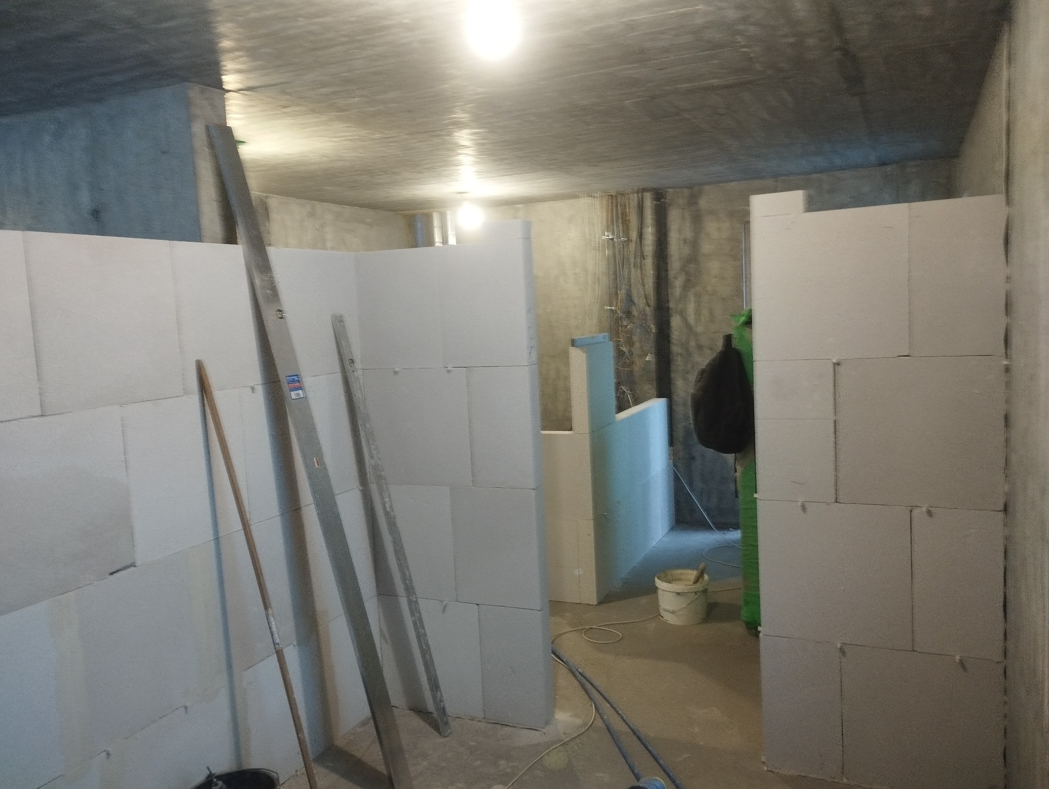 Wnętrze w surowym stanie z wznoszonymi ściankami działowymi z bloczków, widoczne poziomice i wiadro z zaprawą, surowy betonowy strop i ściany w tle.