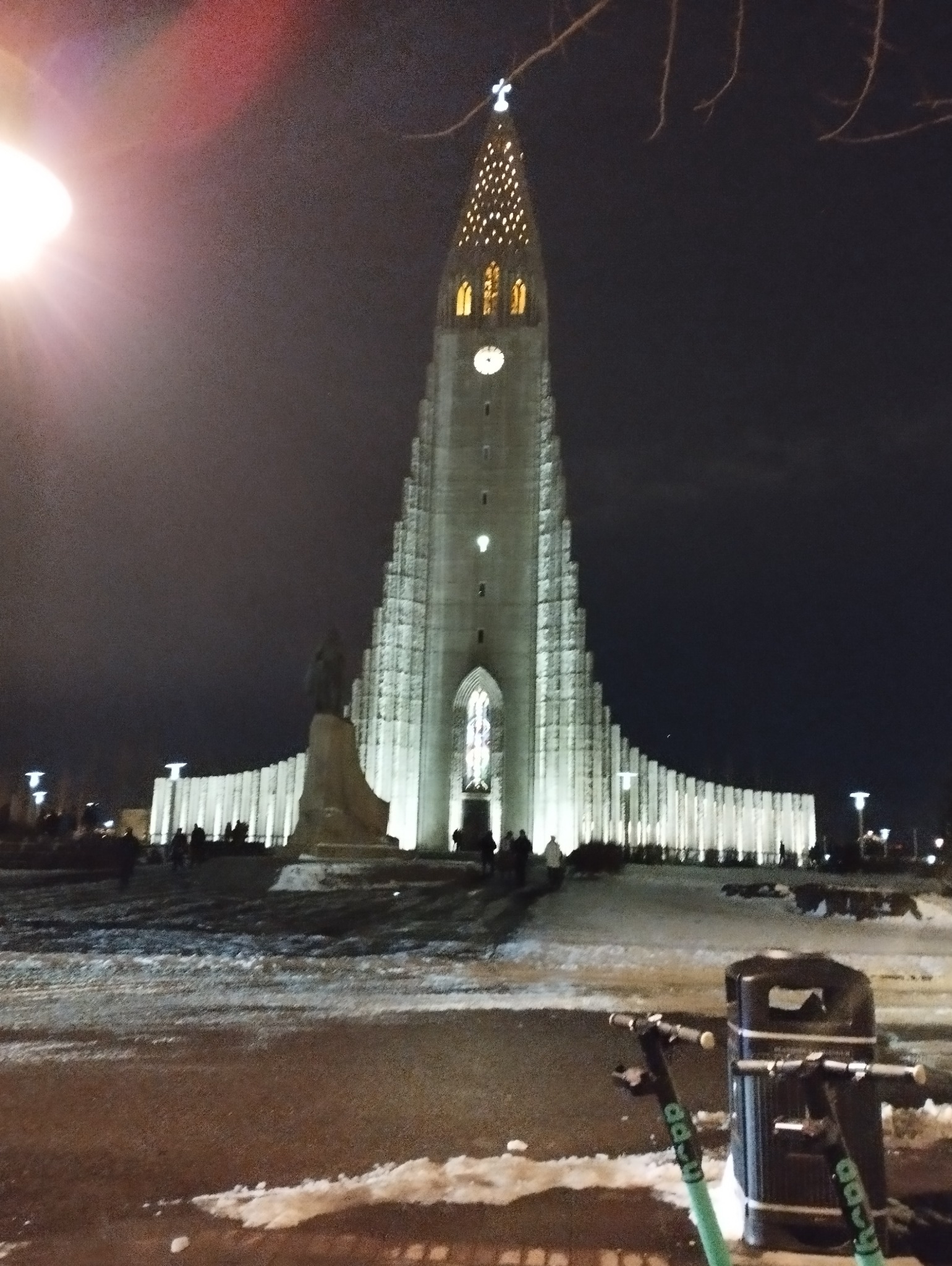 Oświetlony nocą kościół Hallgrímskirkja w Reykjaviku, Islandia, z widocznymi skuterami elektrycznymi na pierwszym planie i śniegiem na ziemi.