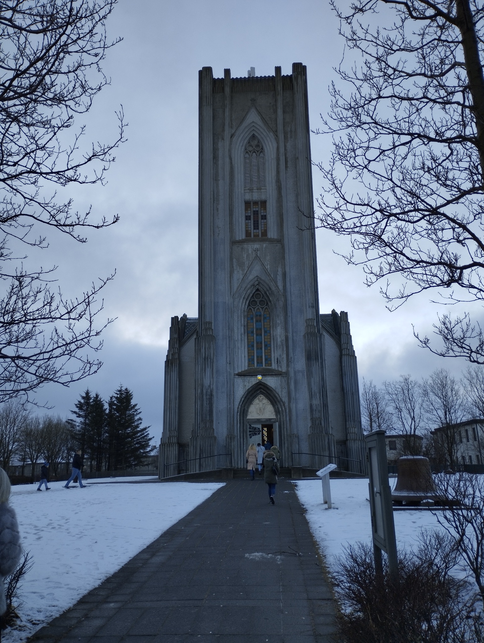 Monumentalna, betonowa konstrukcja sakralna z wysoką wieżą i gotyckimi oknami, widziana z dołu w zimowej scenerii z ludźmi wchodzącymi do środka.
