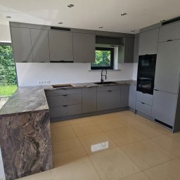 KAMILA MOSPINEK - MEBLE KONCEPT - Szara kuchnia na wymiar z blatem imitującym marmur, z oknem i zabudowaną piekarnią, widok na zewnątrz przez duże okno.