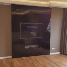 Salon z drewnianą podłogą, ściany w odcieniach brązu, czarna ściana z telewizorem i oświetleniem punktowym, okno z szarymi zasłonami oraz szary szezlong.