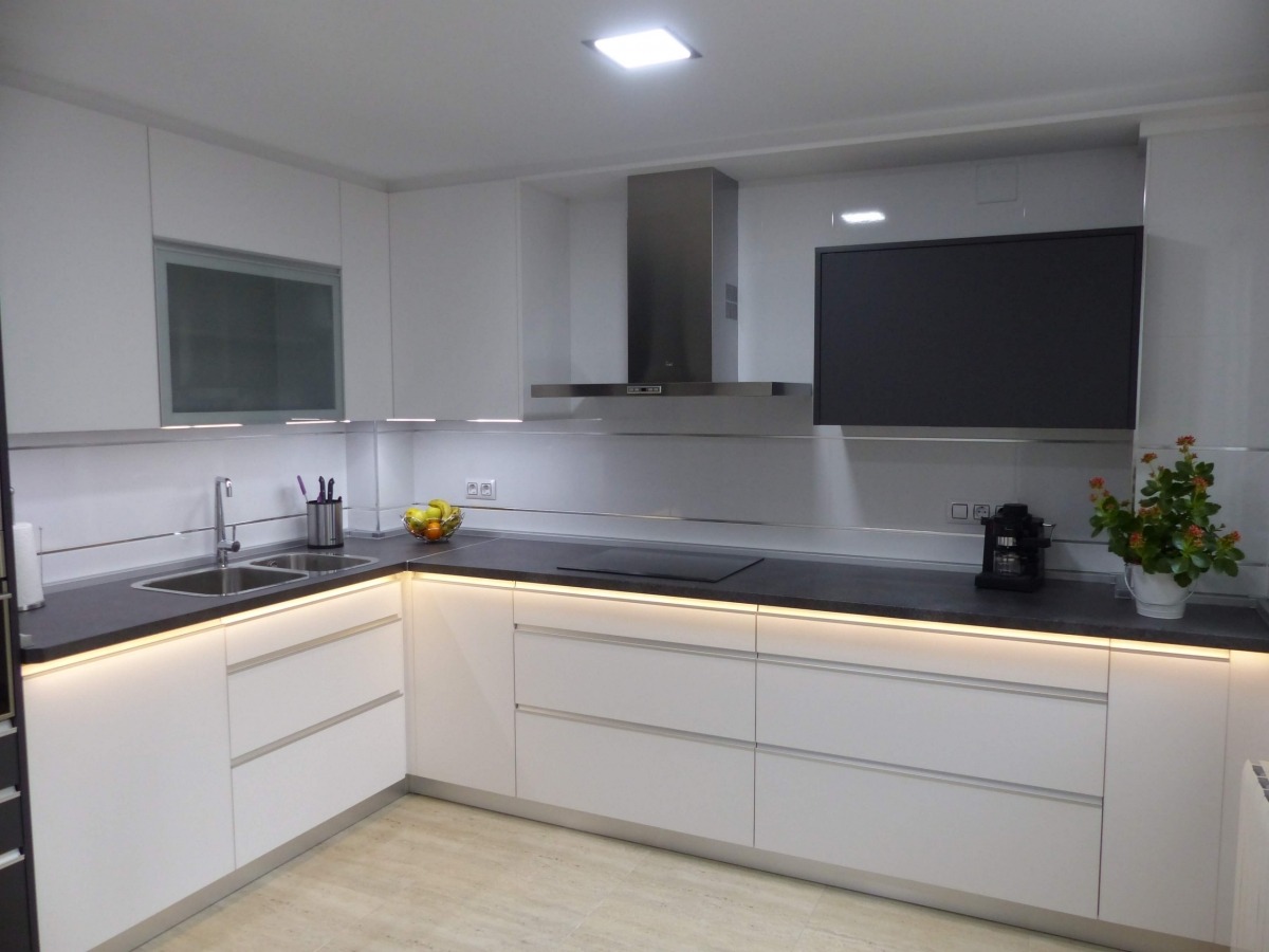 Nowoczesna, biała kuchnia z szarym blatem, oświetleniem LED pod szafkami i srebrnymi akcentami. Minimalistyczny design z kwiatami i ekspresem na blacie.