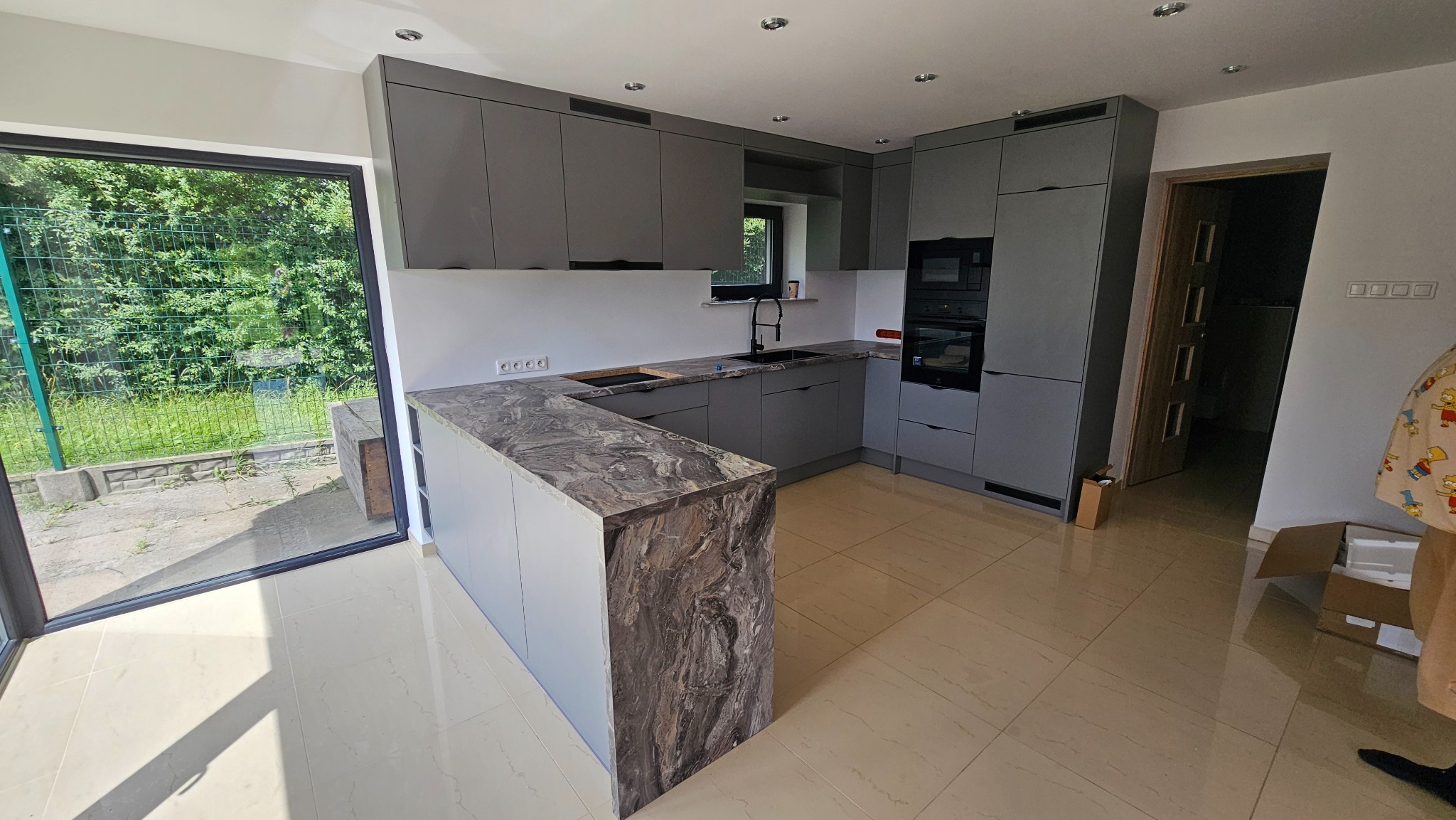 Szara kuchnia na wymiar z marmurowym blatem. Widok na zewnątrz przez duże okno. Nowoczesny design, minimalistyczne fronty, oświetlenie punktowe.