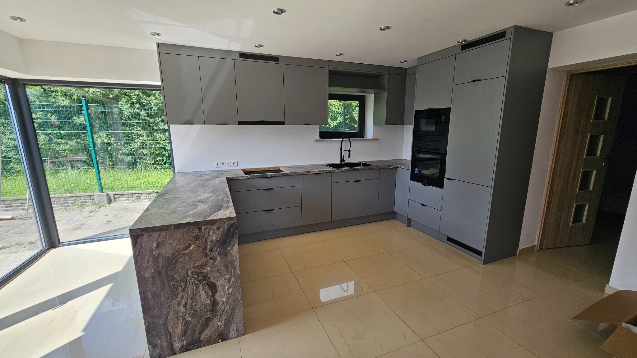 Szara kuchnia na wymiar z blatem imitującym marmur, z oknem i zabudowaną piekarnią, widok na zewnątrz przez duże okno.