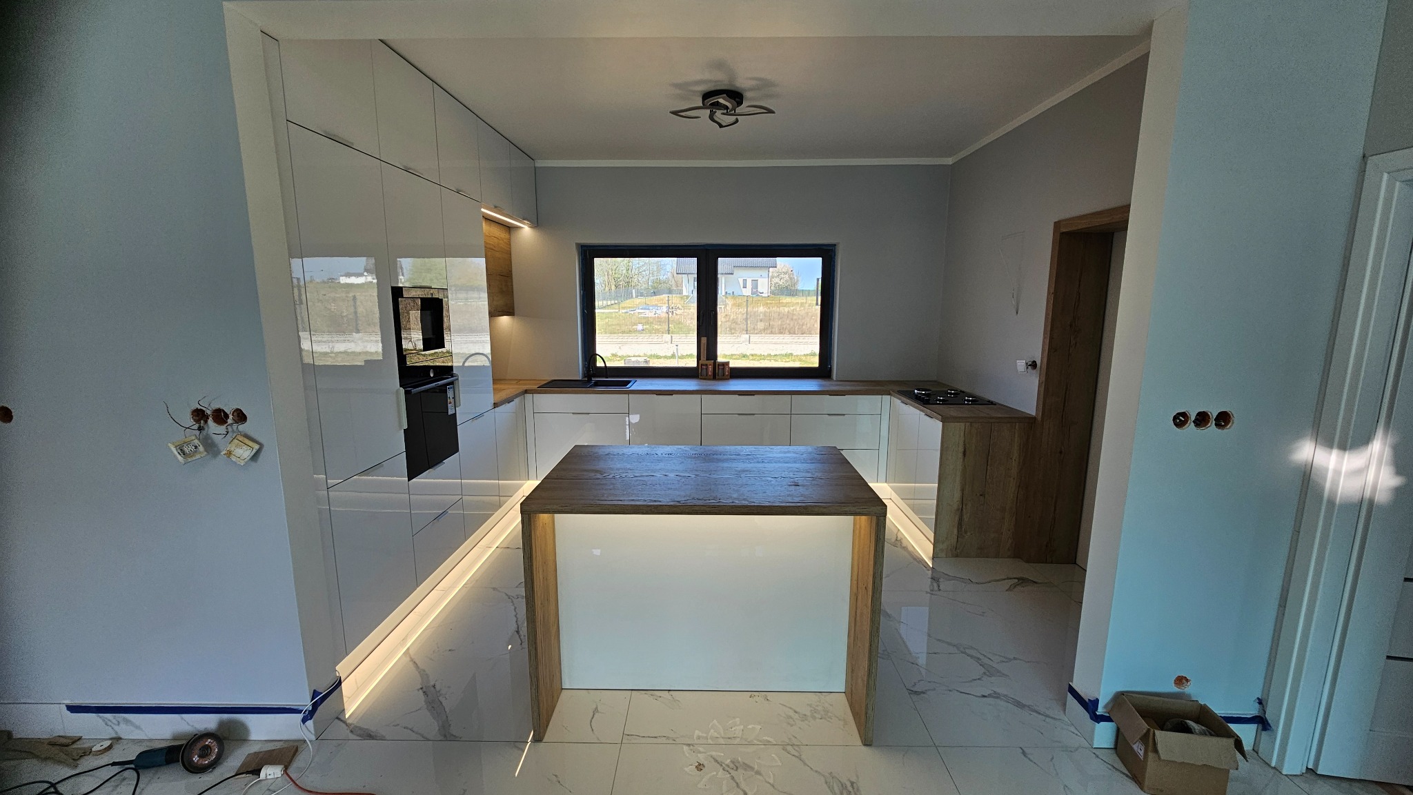 Nowoczesna kuchnia z wyspą i białymi, lakierowanymi frontami. Blat drewniany, oświetlenie LED pod szafkami. Widoczne gniazdka elektryczne na ścianie.