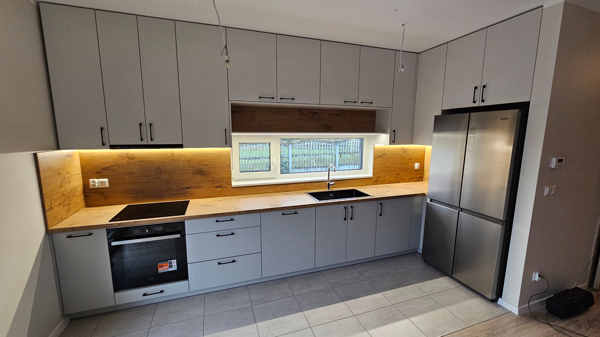 Jasna kuchnia z drewnianym blatem i podświetleniem LED, czarne uchwyty, duża lodówka. Nowoczesne wykończenie, okno z widokiem na zieleń.