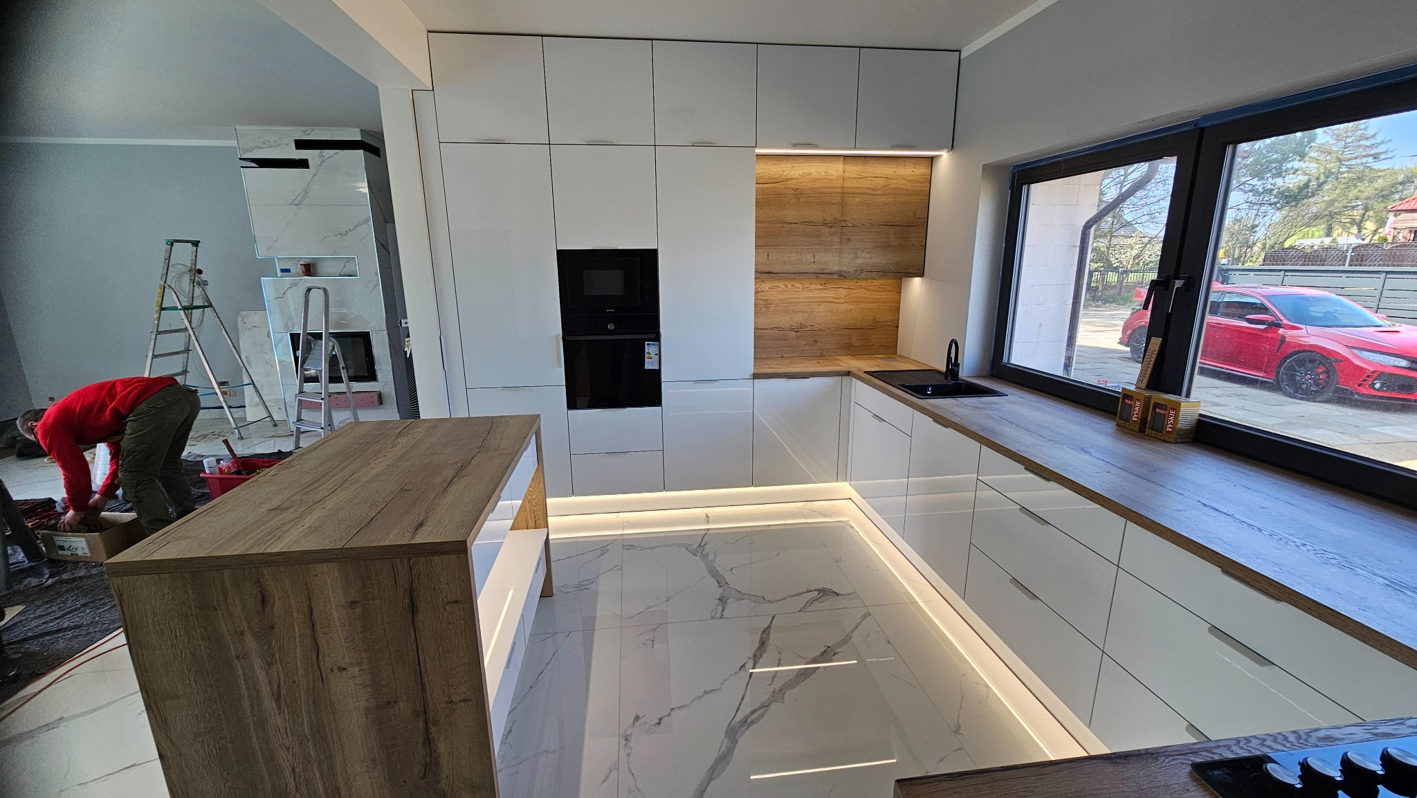 Montaż nowoczesnej kuchni: białe, lakierowane fronty, drewniany blat i wyspa, oświetlenie LED pod szafkami i na podłodze. W tle widoczny monter i fragment salonu.