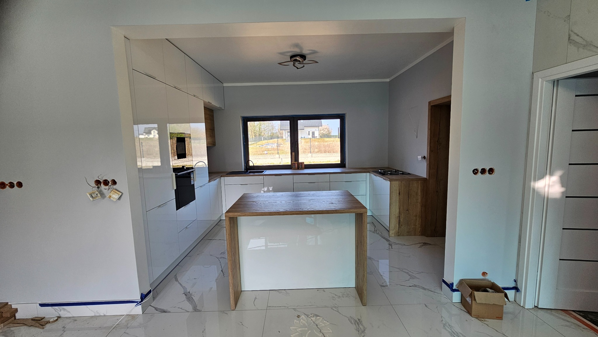 Nowoczesna kuchnia z wyspą i białymi frontami na wysoki połysk, drewniany blat, okno z widokiem na dom. Częściowo widoczne instalacje elektryczne.