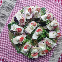 Dekoracyjna taca z kanapkami: pieczywo, szynka, ser, sałata, pomidory, sos i kiełki, na tle czerwono-białej kraty z serduszkami i jutowej podkładki.