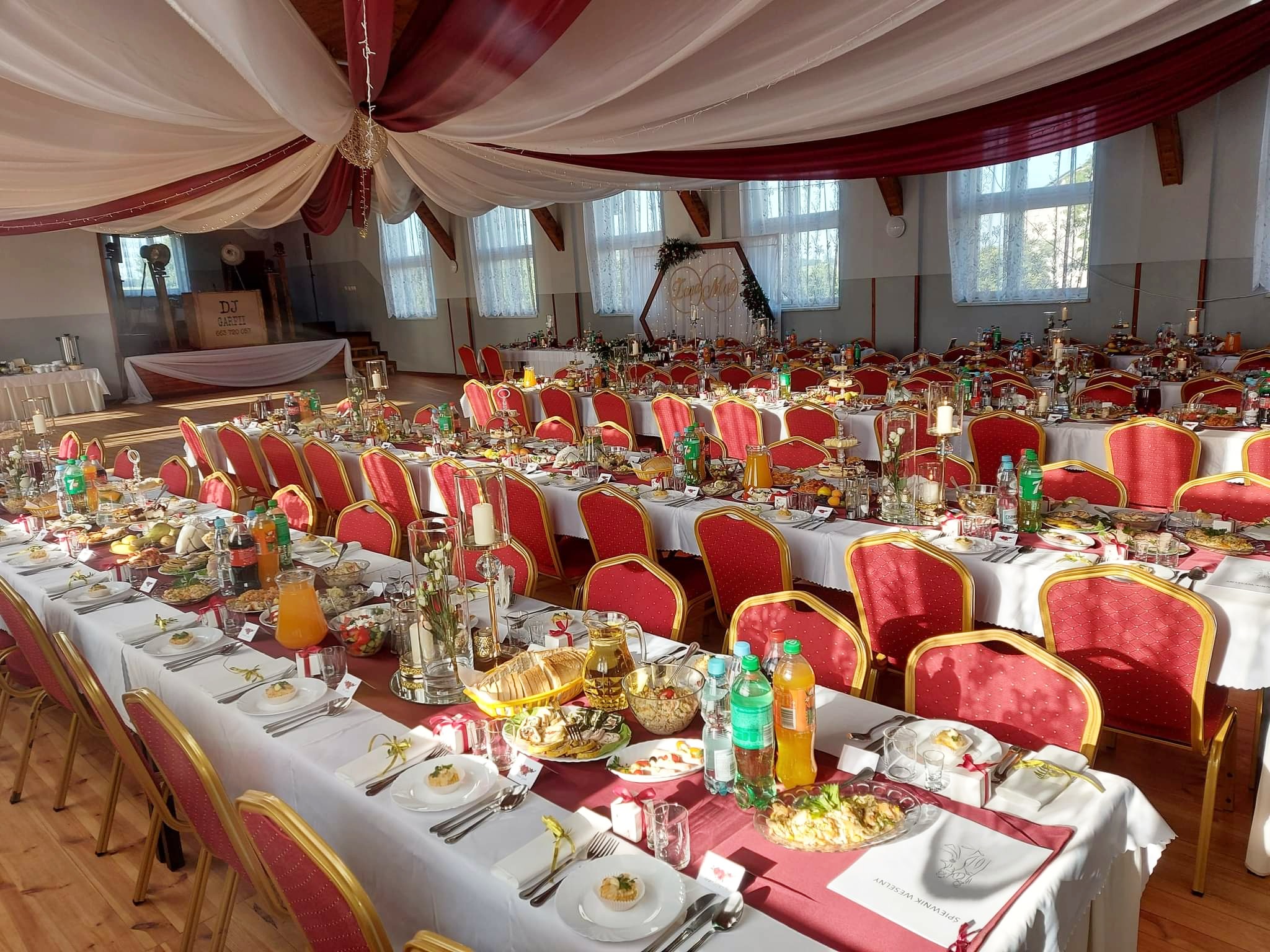 Sala bankietowa z długimi stołami nakrytymi białymi obrusami i czerwonymi bieżnikami, czerwone krzesła, dekoracje sufitowe z białych i czerwonych materiałów, na stołach zastawa, potrawy, napoje...