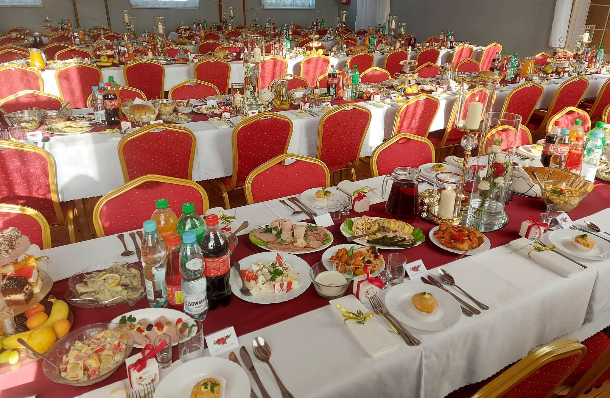 Sala bankietowa z długimi stołami nakrytymi białymi obrusami z bordowymi bieżnikami, zastawiona talerzami z przekąskami, napojami, dekoracjami kwiatowymi i świecznikami, krzesła z czerwonym obiciem.