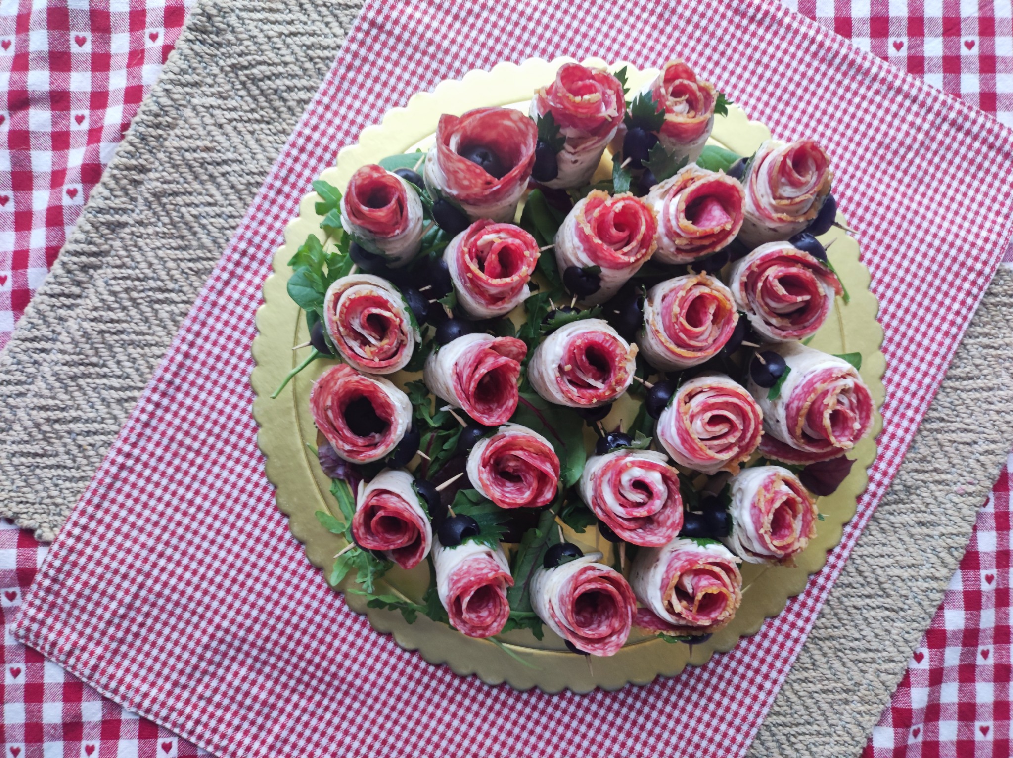 Dekoracyjna kompozycja z roladek salami ułożonych w kształt kwiatów na złotej tacy, ozdobiona rukolą i oliwkami, na tle czerwonej kraty i beżowej plecionki.