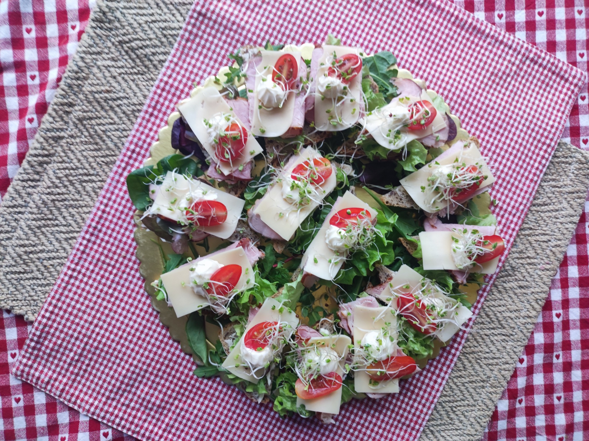 Dekoracyjna taca z kanapkami: pieczywo, szynka, ser, sałata, pomidory, sos i kiełki, na tle czerwono-białej kraty z serduszkami i jutowej podkładki.