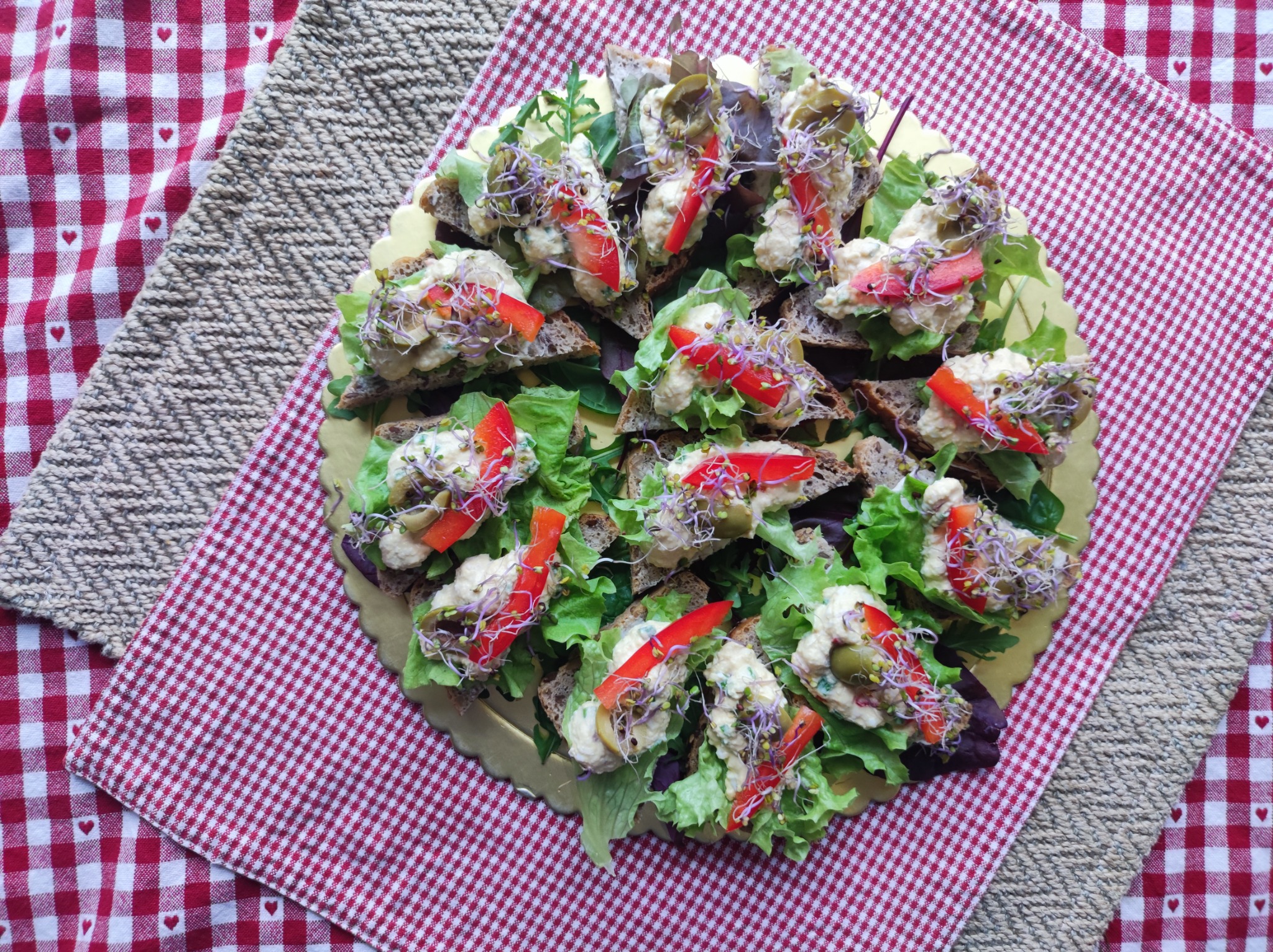 Apetyczne kanapeczki na ciemnym pieczywie z sałatą, pastą, czerwoną papryką i kiełkami, ułożone na złotej tacy na tle czerwono-białej i beżowej tkaniny.