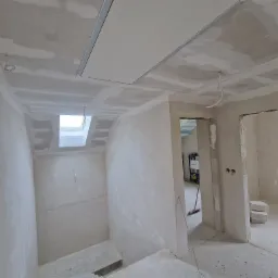 Wnętrze w trakcie remontu: ściany pokryte szpachlą, widoczne płyty gipsowo-kartonowe na suficie, instalacje elektryczne wystające ze ściany, surowe wykończenie schodów i widoczne elementy...