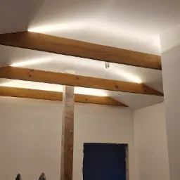Drewniane belki stropowe w poddaszu z ukrytym oświetleniem LED, filar zabezpieczony folią, otwory w ścianach przygotowane do dalszych prac wykończeniowych.