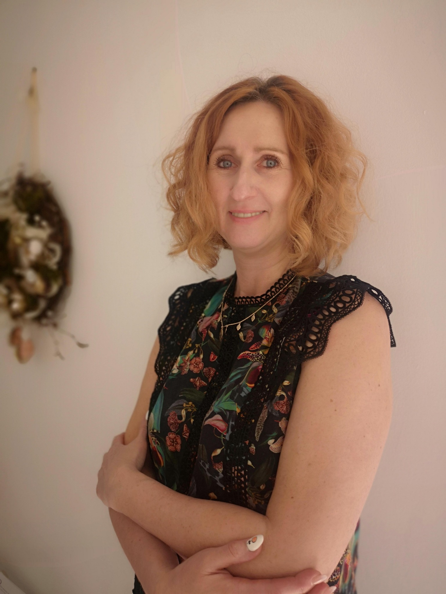 Portret uśmiechniętej kobiety o rudych włosach, stojącej na tle jasnej ściany z dekoracją z suszonych roślin.