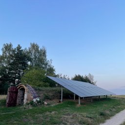 Holidom Przemysław Brzóska - Panel słoneczny zamontowany na łące obok ziemianki z otwartymi, drewnianymi drzwiami w letni wieczór.