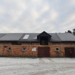 Holidom Przemysław Brzóska - Instalacja paneli słonecznych na dachu ceglanego budynku gospodarczego z widocznym oknem dachowym i osobą w czerwonej kamizelce przy drzwiach, zimowy krajobraz w tle.