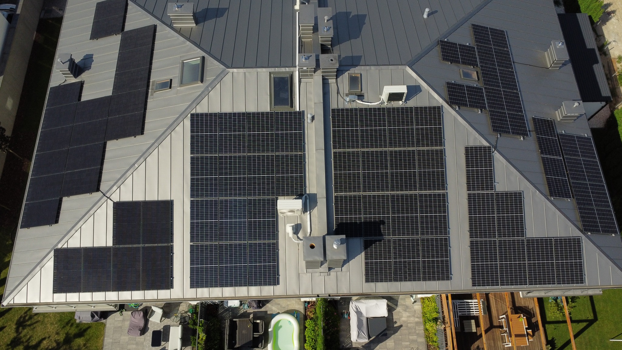 Widok z góry na szary dach z panelami słonecznymi, klimatyzatorami i oknami dachowymi; poniżej widoczny ogród z basenem i meblami ogrodowymi.