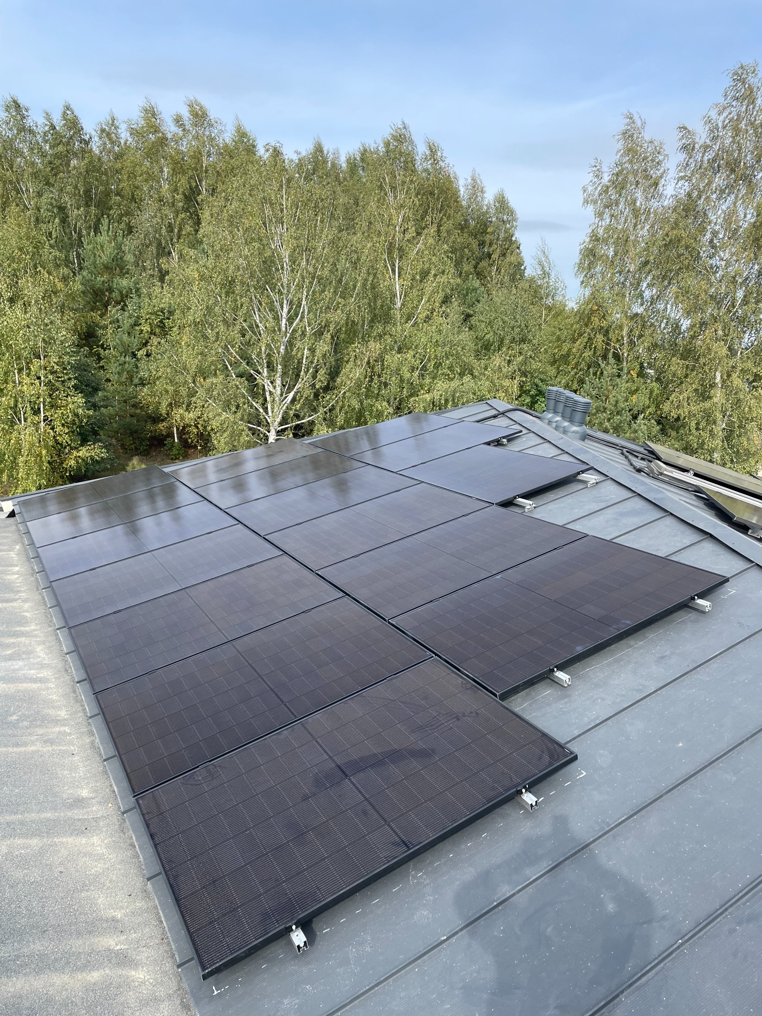 Częściowo zamontowane panele słoneczne na szarym dachu z blachy na tle drzew i błękitnego nieba.