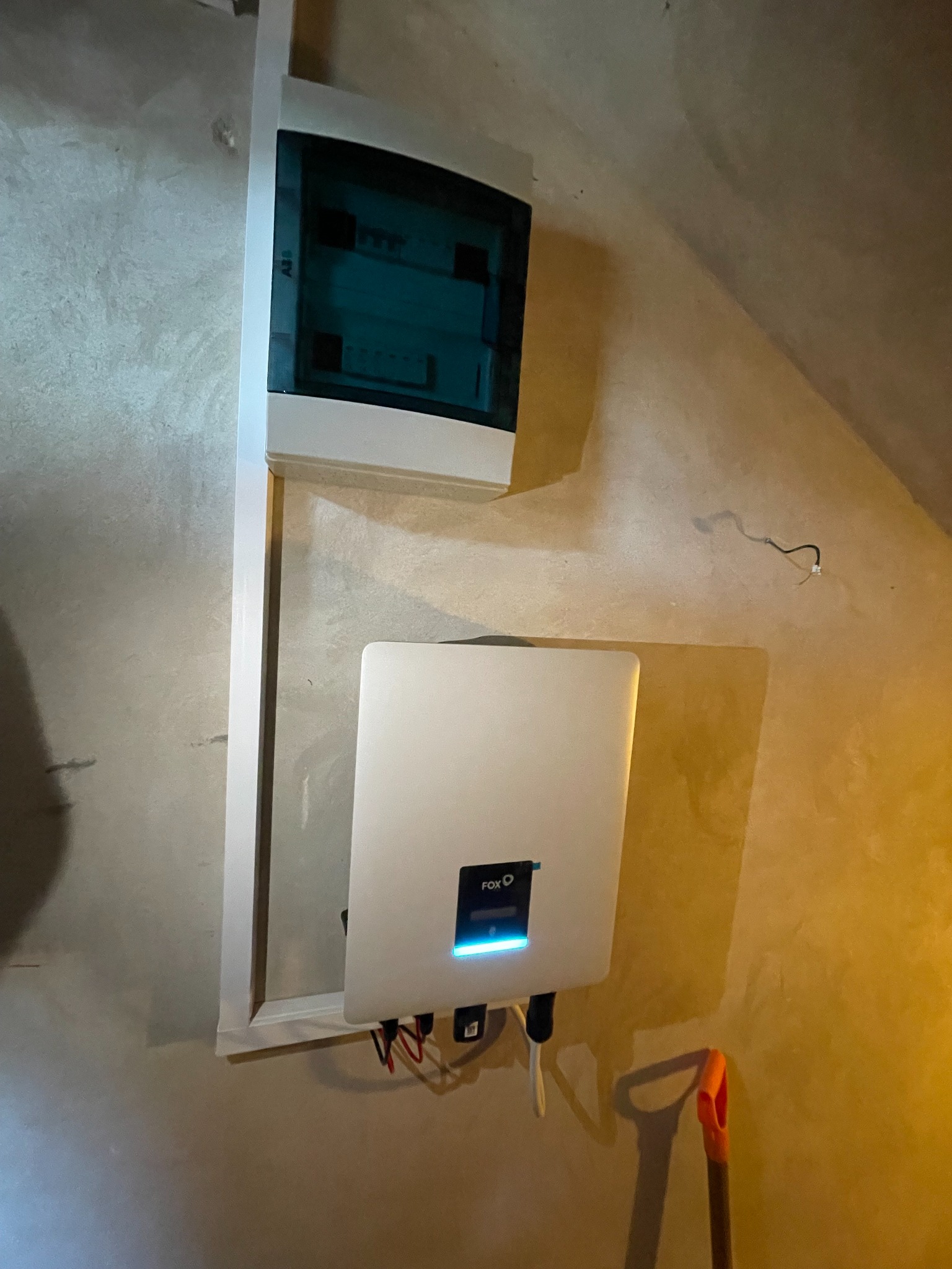Biała skrzynka rozdzielcza i falownik marki FOX na ścianie w surowym wnętrzu, widoczne przewody i fragment łopaty.