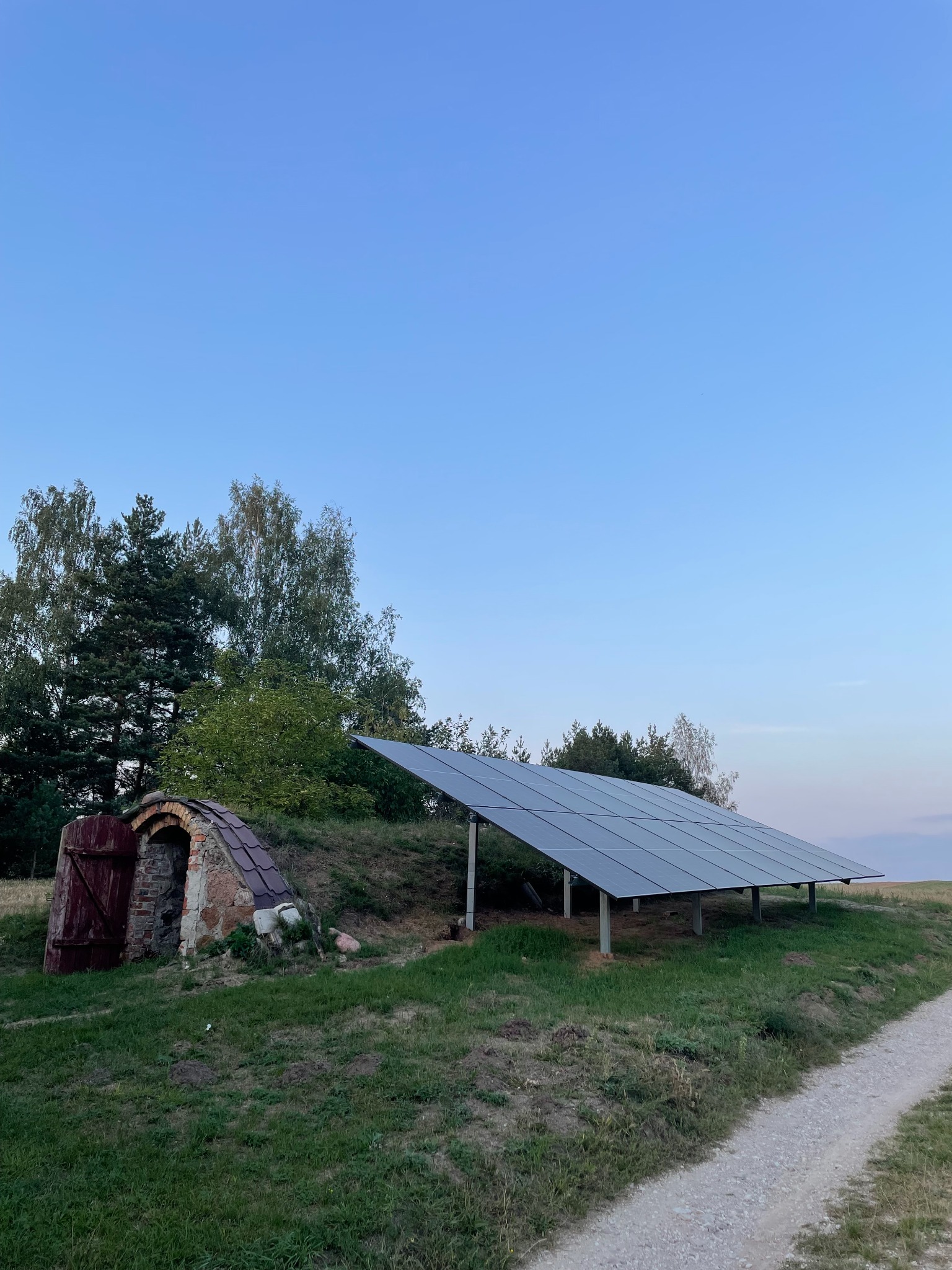 Panel słoneczny zamontowany na łące obok ziemianki z otwartymi, drewnianymi drzwiami w letni wieczór.