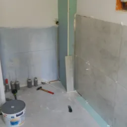 Wnętrze łazienki w trakcie remontu: częściowo wyłożone szarymi płytkami, widoczne narzędzia, poziomica, wiaderko z farbą i niedokończone ściany.