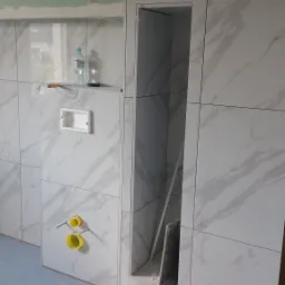 Wykończenie łazienki w trakcie remontu: białe płytki imitujące marmur na ścianach, wnęka w ścianie, żółte elementy instalacji wodnej, butelki z wodą na półce i surowy stan podłogi.