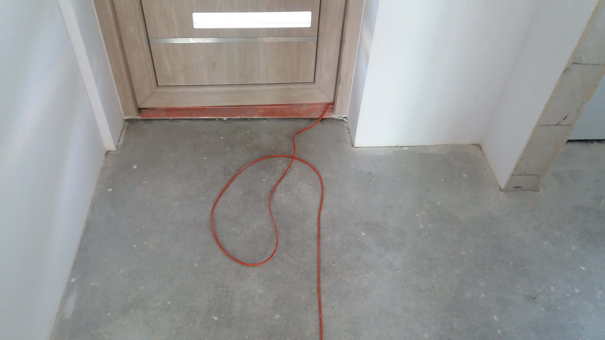 Surowy betonowy podkład podłogowy w trakcie prac wykończeniowych, widoczna pomarańczowa linka i fragment ościeżnicy drzwiowej z drewnopodobną okleiną.