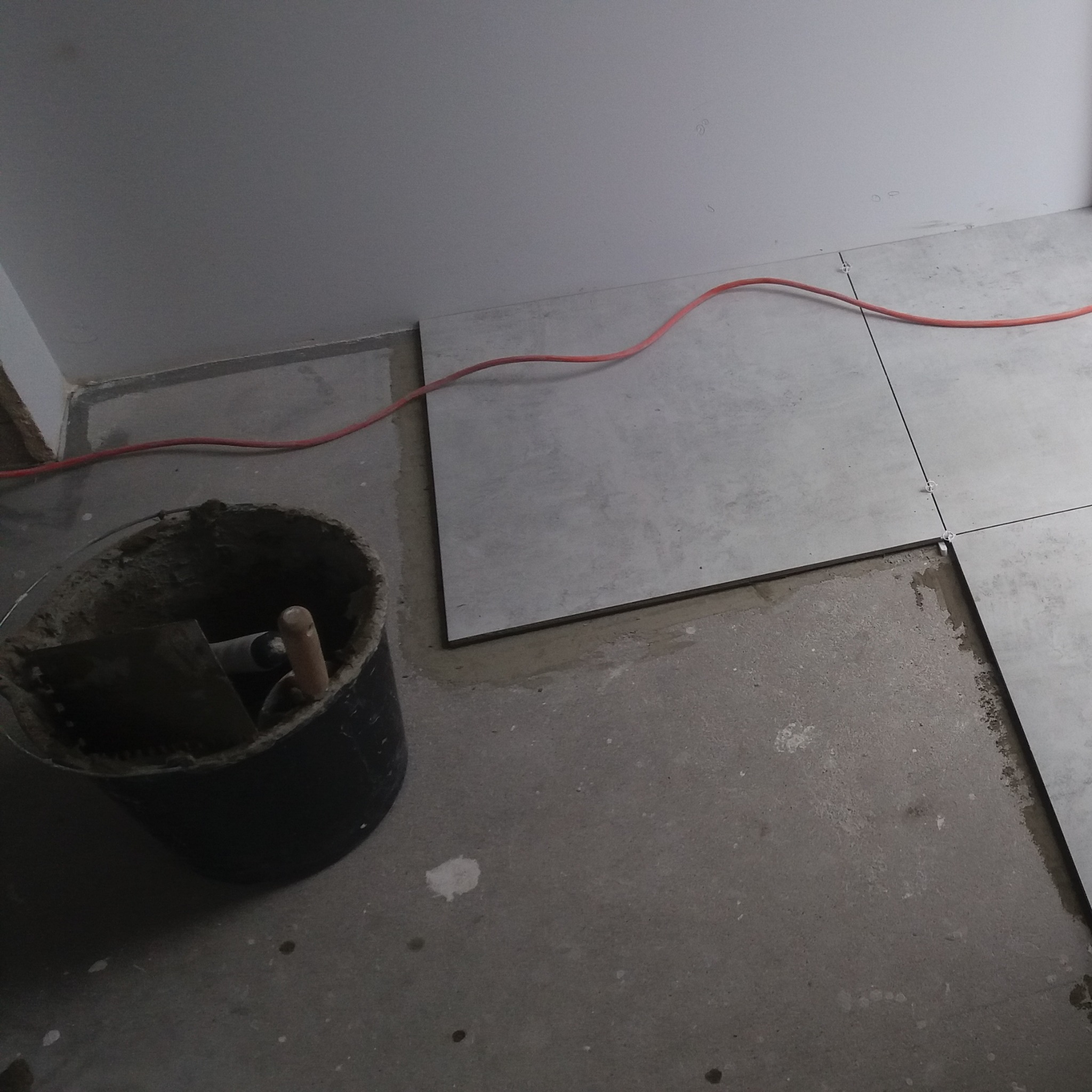 Układanie jasnoszarych płytek podłogowych w pomieszczeniu, widoczny kubeł z zaprawą i narzędziami oraz czerwony kabel na podłodze.
