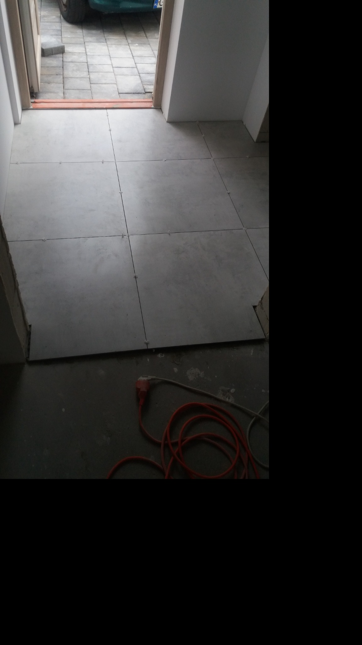 Szare płytki podłogowe, ułożone w kwadraty z plastikowymi krzyżykami dystansowymi, w trakcie montażu, widoczne kable zasilające na podłodze.