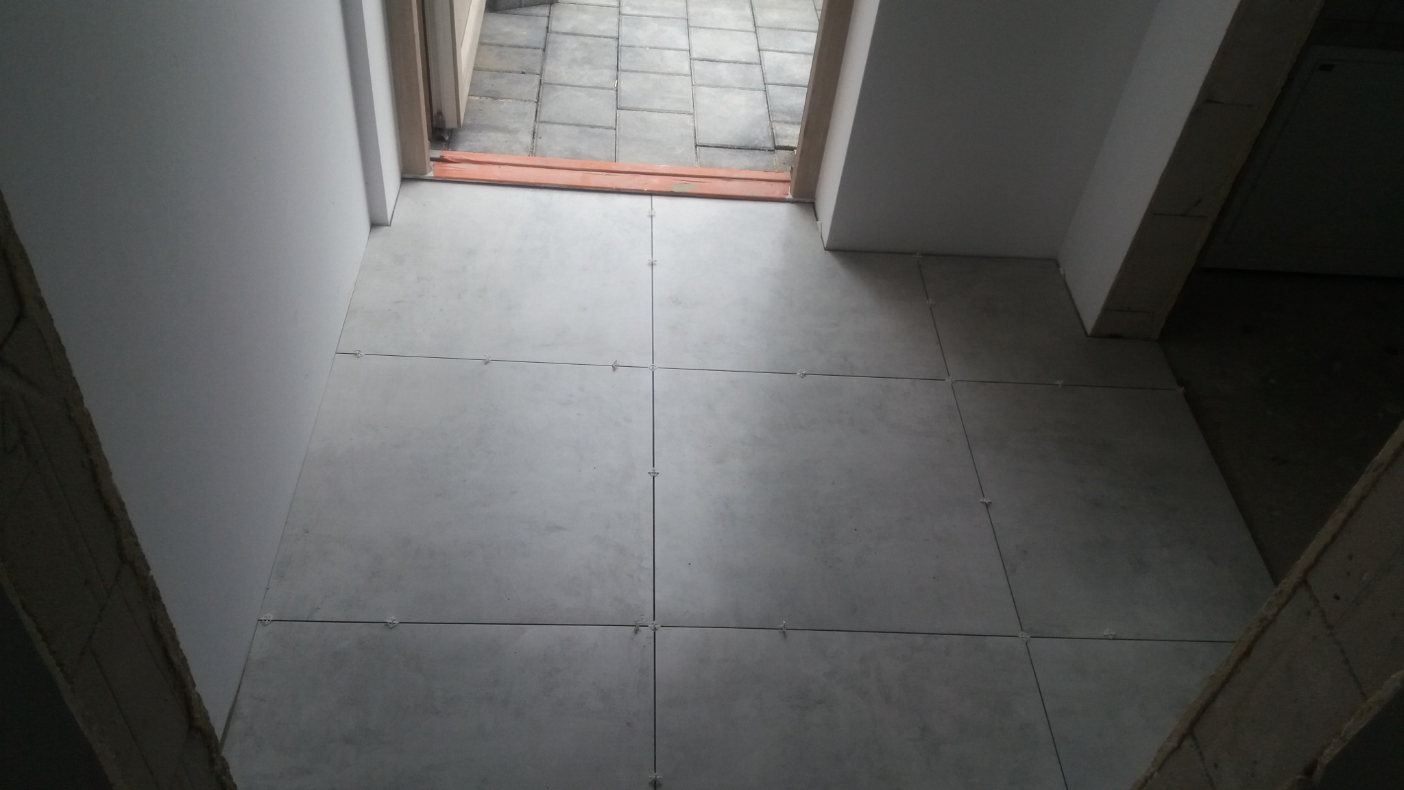 Świeżo ułożone szare płytki podłogowe z krzyżykami dystansowymi, widok z góry na próg drzwiowy i fragment kostki brukowej na zewnątrz.