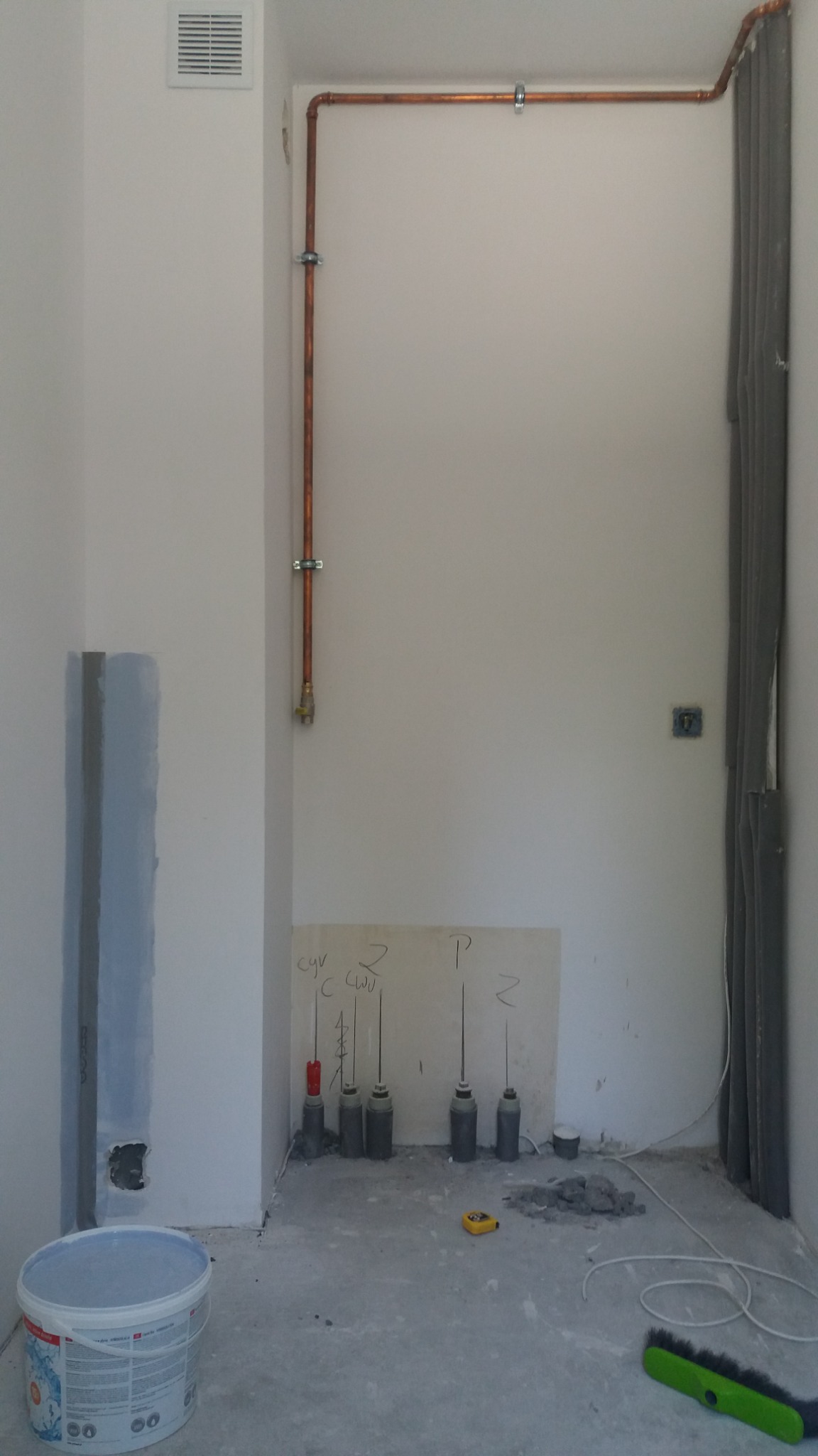 Przygotowane stanowisko pracy w pomieszczeniu w trakcie remontu: miedziane rury przy ścianie, zaprawa, poziomica, wiadro farby i szczotka do zamiatania.