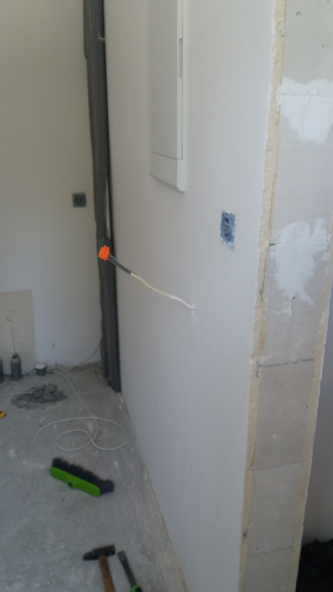 Wnętrze w trakcie remontu: ściana po szpachlowaniu z wystającym przewodem elektrycznym, widoczna puszka instalacyjna, fragment ościeżnicy, na podłodze szczotka i młotek.