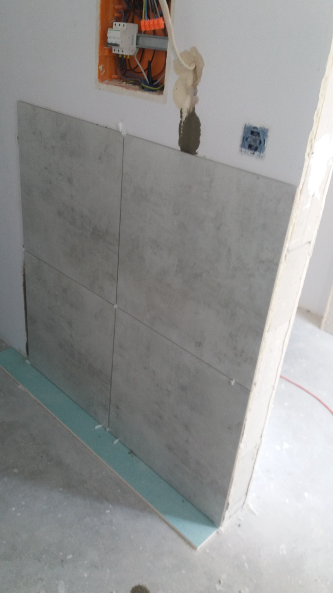 Montaż szarych płytek ceramicznych na ścianie w trakcie remontu, widoczne krzyżyki dystansowe, puszka elektryczna z przewodami i obudowa gniazdka.