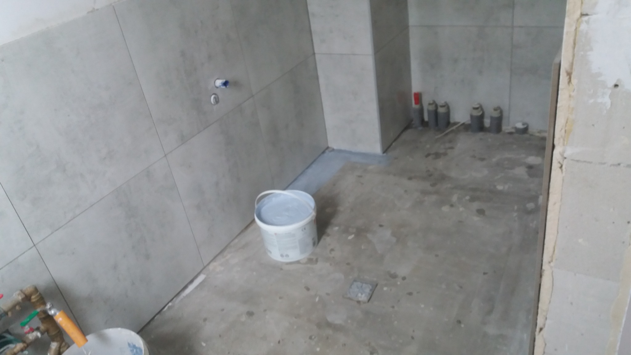 Świeżo wykafelkowana łazienka w trakcie remontu, widoczny biały pojemnik z zaprawą, fragment instalacji wodnej i odpływów kanalizacyjnych.