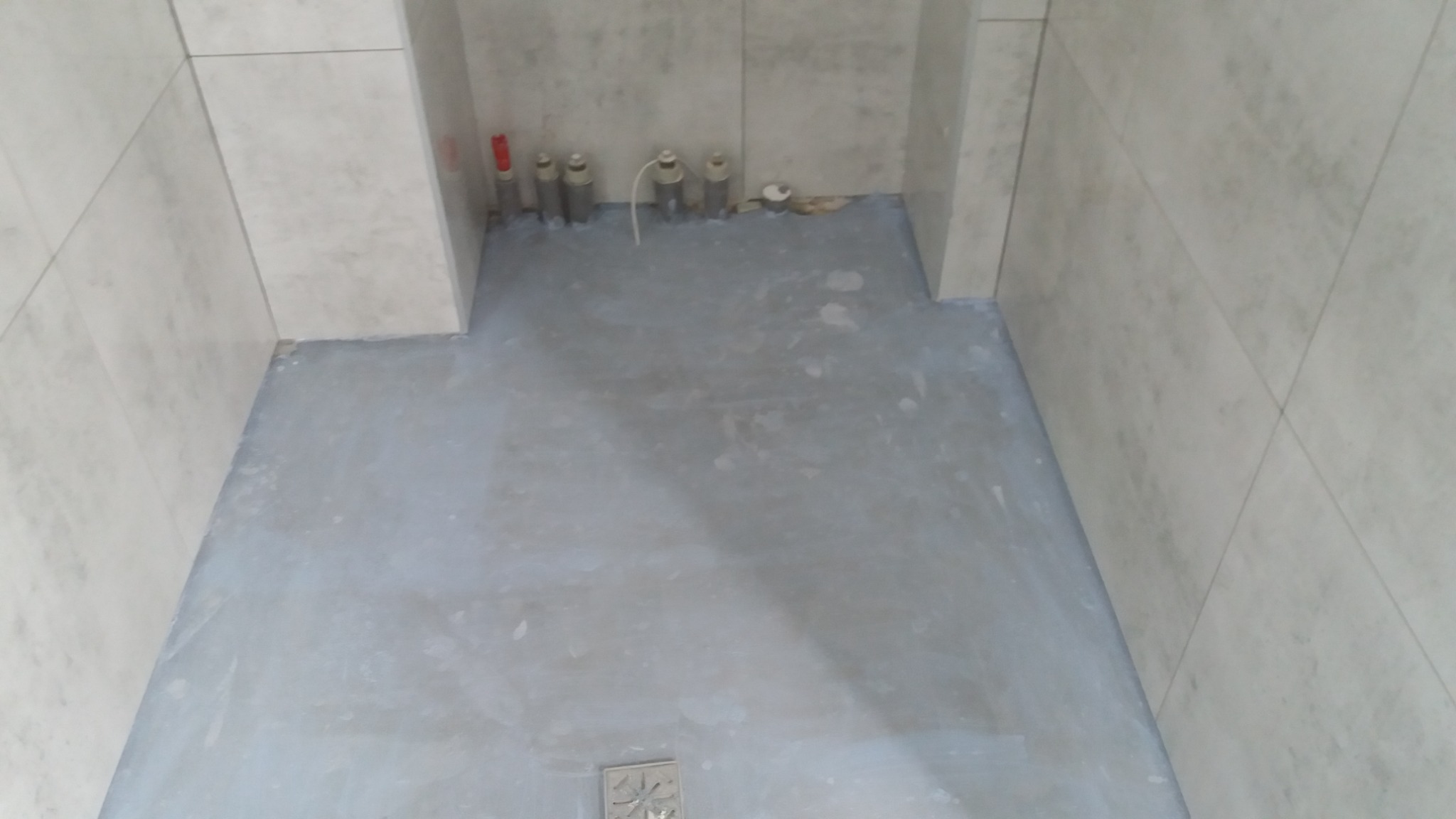 Przygotowana podłoga w kabinie prysznicowej z nałożoną warstwą hydroizolacji, widoczne rury instalacji wodnej przy ścianie wyłożonej płytkami.