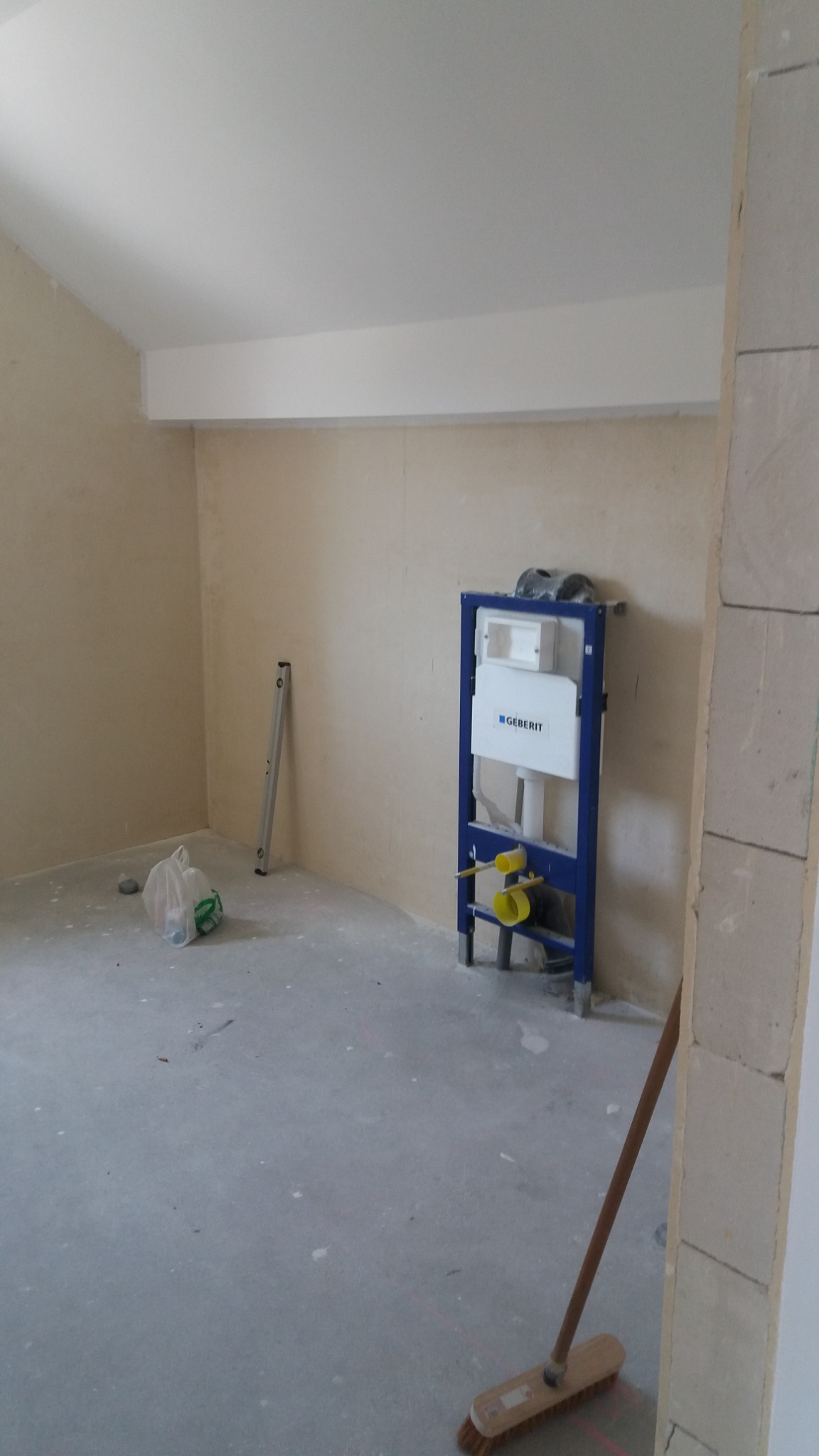 Wnętrze łazienki w trakcie remontu, z zamontowanym stelażem podtynkowym Geberit, poziomica oparta o ścianę, widoczny surowy stan ścian i podłogi, oraz częściowo wymurowana ściana z bloczków.