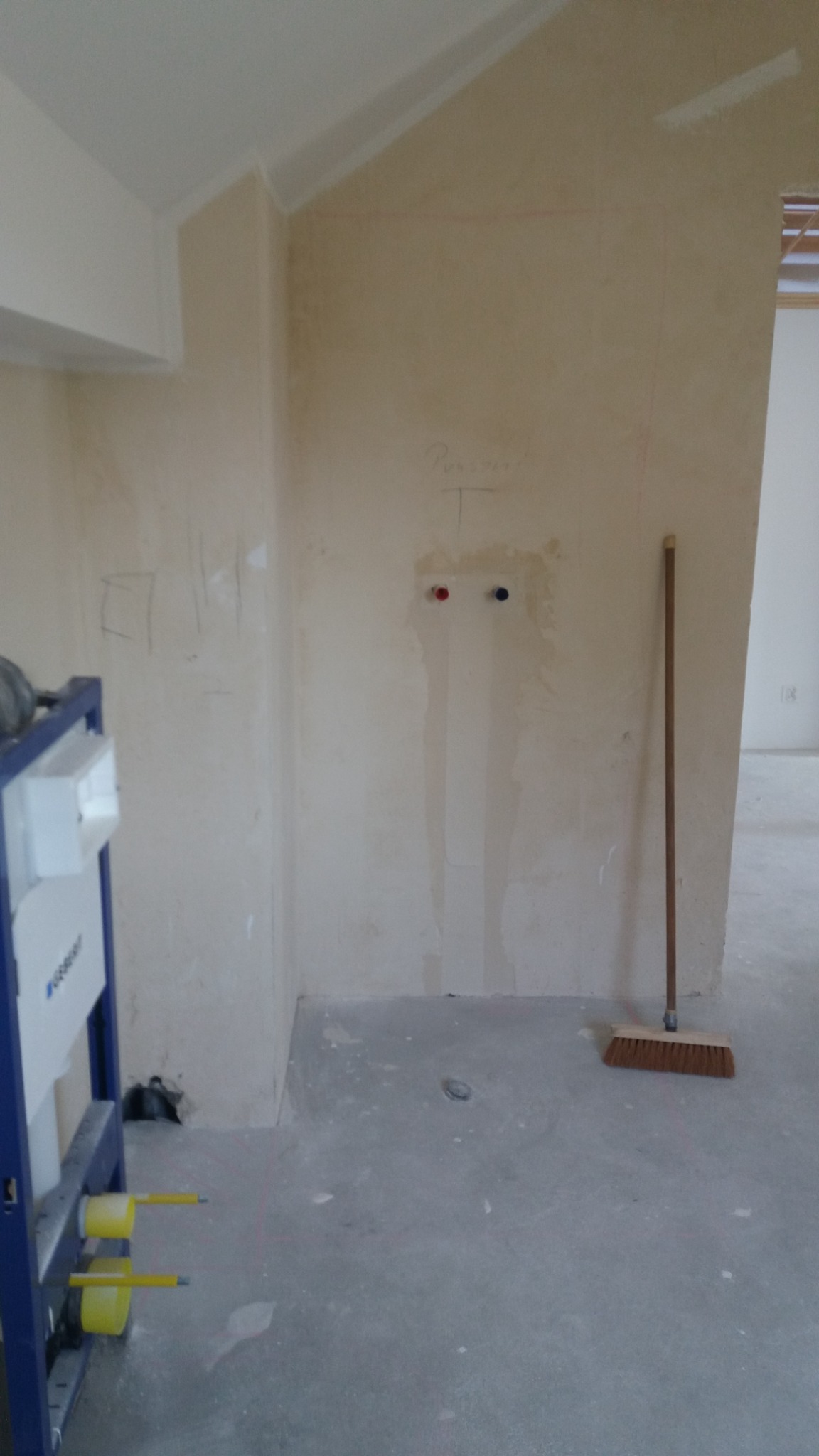 Niedokończona łazienka z podtynkowym stelażem WC Geberit, instalacją wodną z niebieskim i czerwonym zaworem, brudną ścianą z odręcznymi notatkami i miotłą opartą o ścianę.