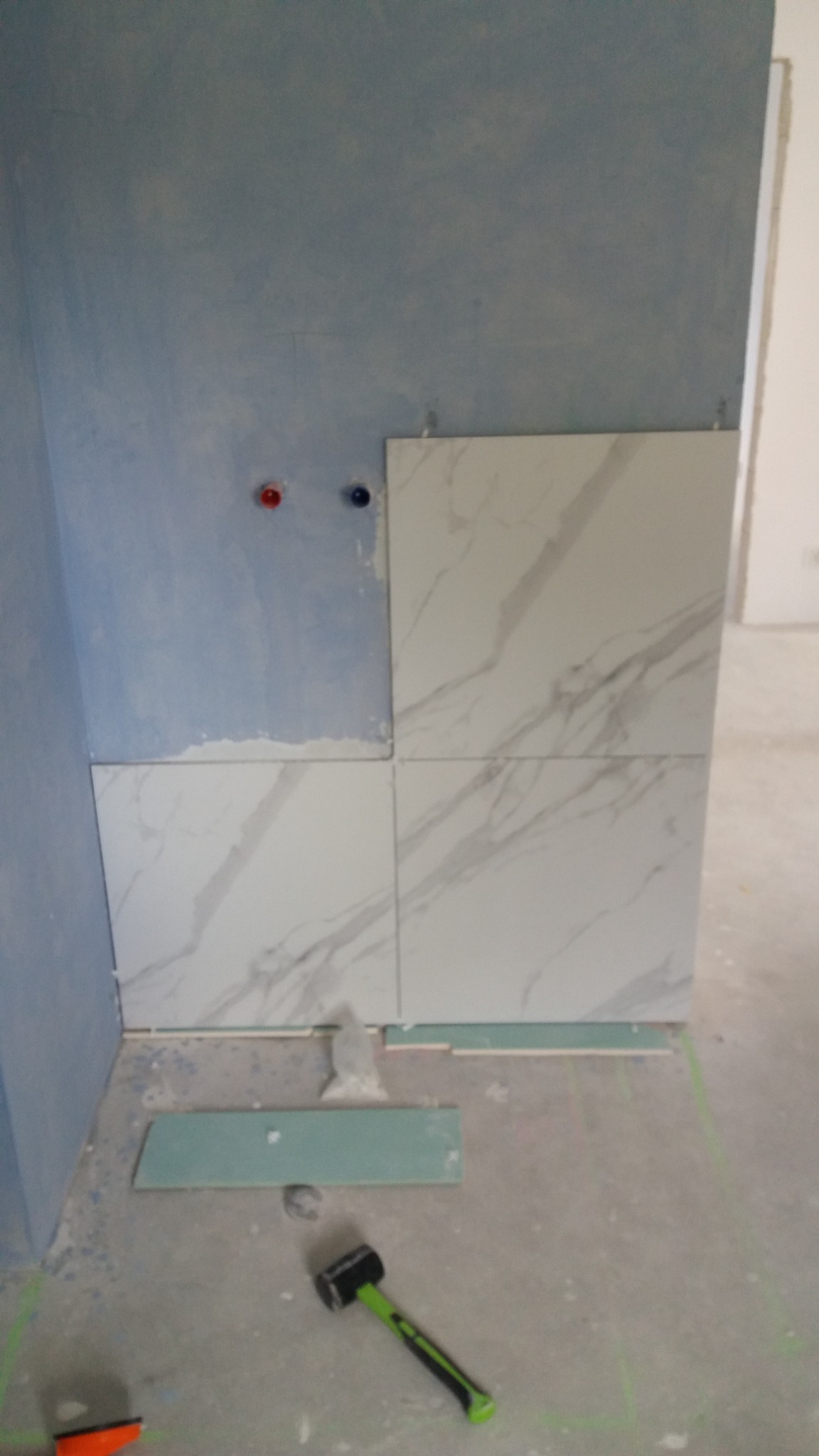 Fragment ściany z częściowo ułożonymi płytkami imitującymi marmur, widoczne przygotowane płytki, poziomica i młotek z zielonym uchwytem.