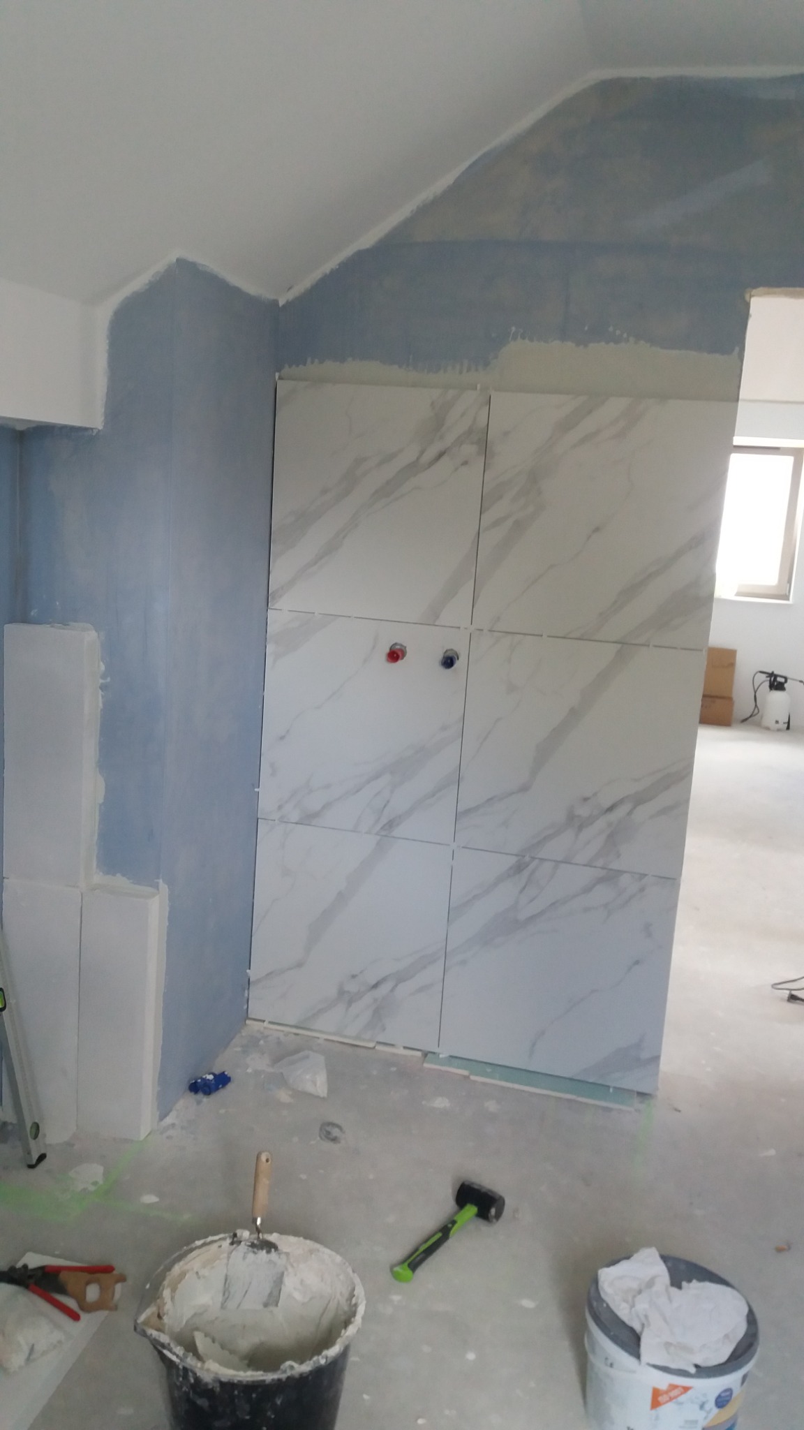 Ściana w trakcie wykańczania z ułożonymi płytkami imitującymi marmur, widoczne przygotowane bloki, wiadro z zaprawą i poziomica laserowa na podłodze w trakcie remontu.