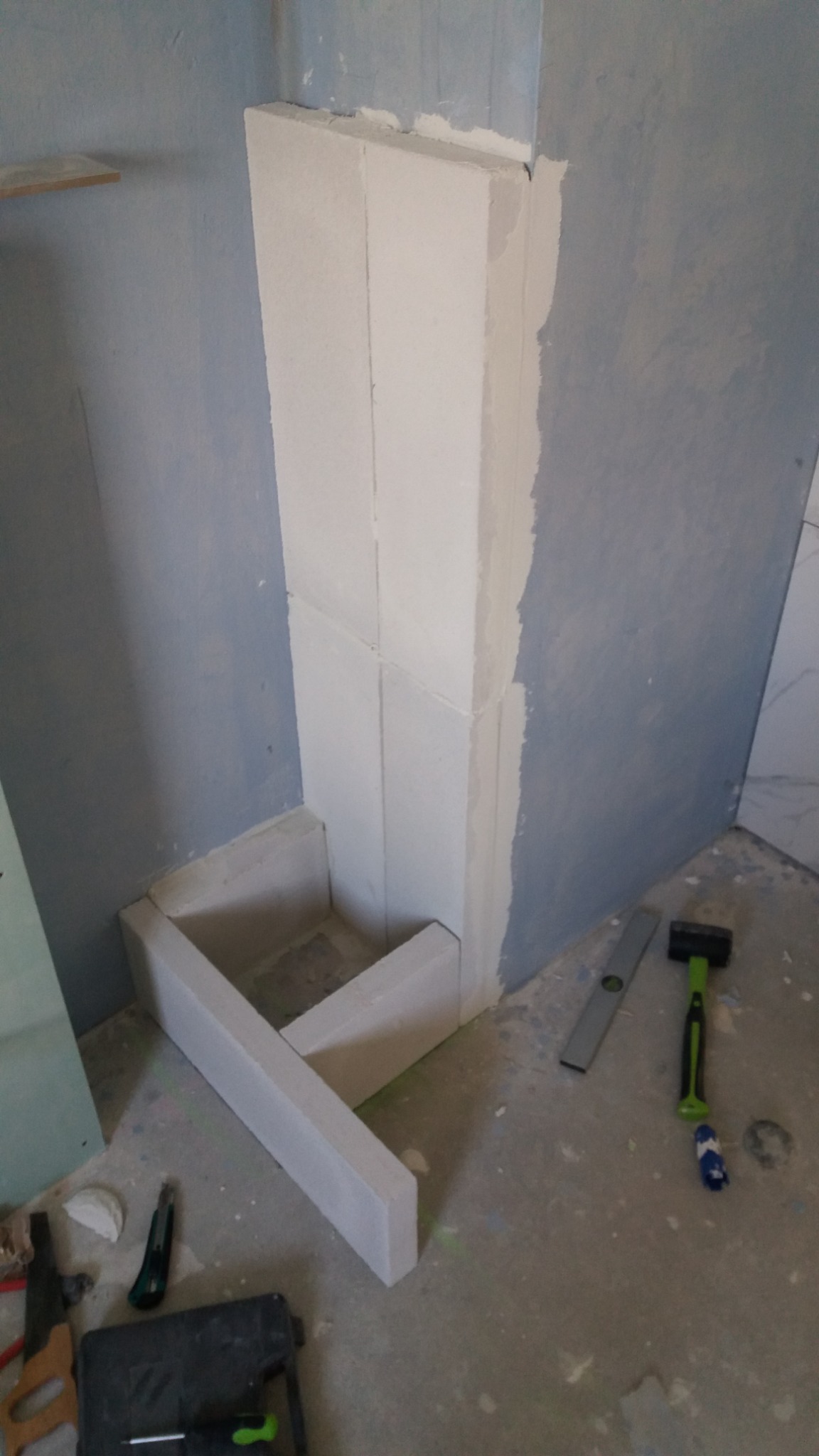 Wznoszenie konstrukcji z białych bloczków, prawdopodobnie obudowa rury, w trakcie prac wykończeniowych w łazience, widoczne narzędzia: poziomica, młotek i skrzynka narzędziowa.