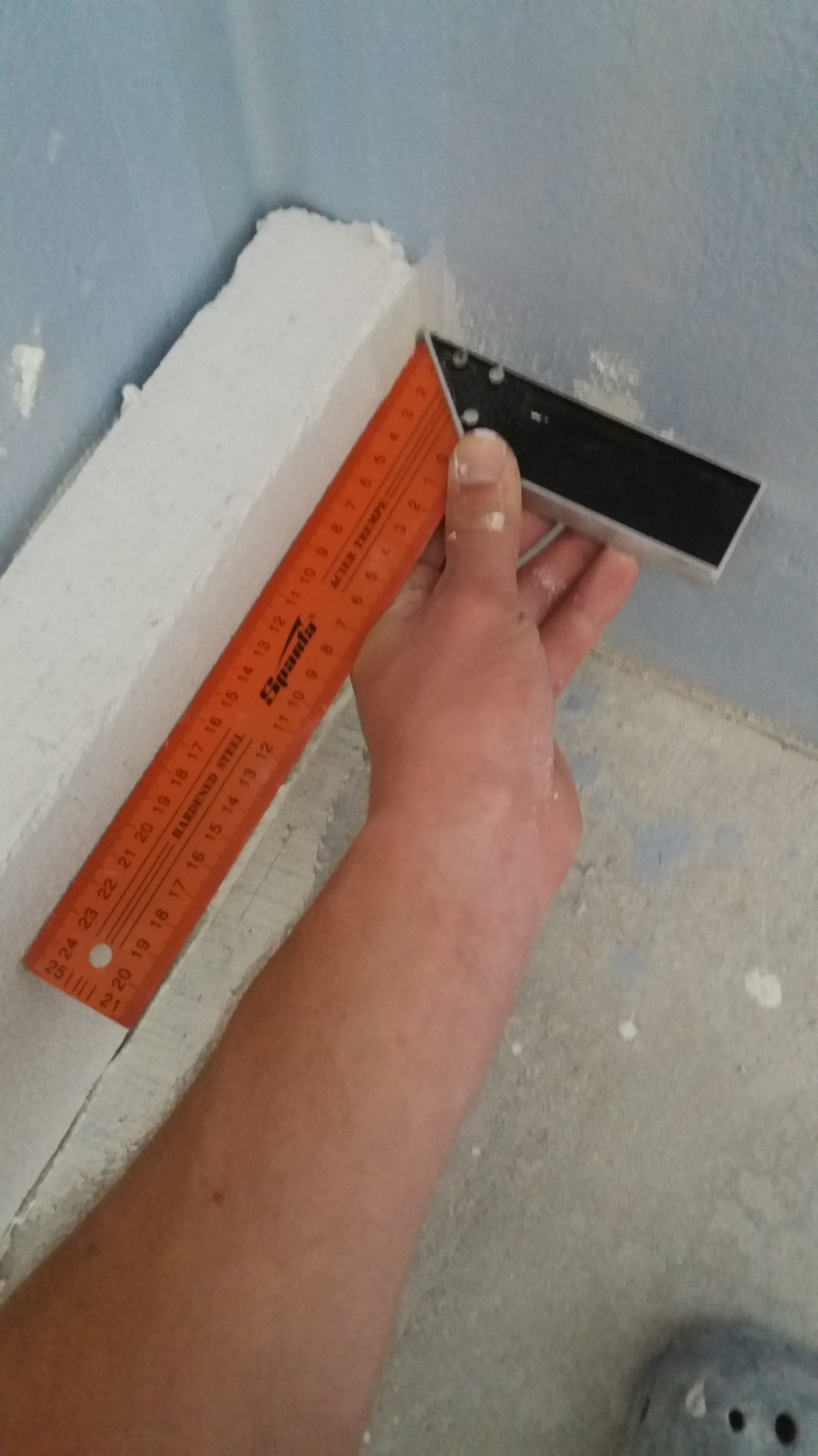 Pomiar kąta prostego w rogu ściany po nałożeniu gładzi, pomarańczowa miarka Spanta z widocznymi numerami, biała gładź na styku niebieskiej ściany i podłogi.