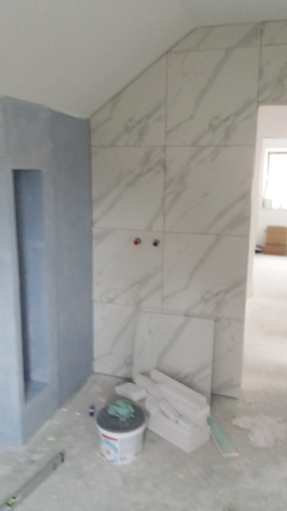 Ściana wyłożona marmurowymi płytkami w łazience pod skosem, z widocznymi otworami na armaturę, obok wiadro z farbą i zapas płytek.