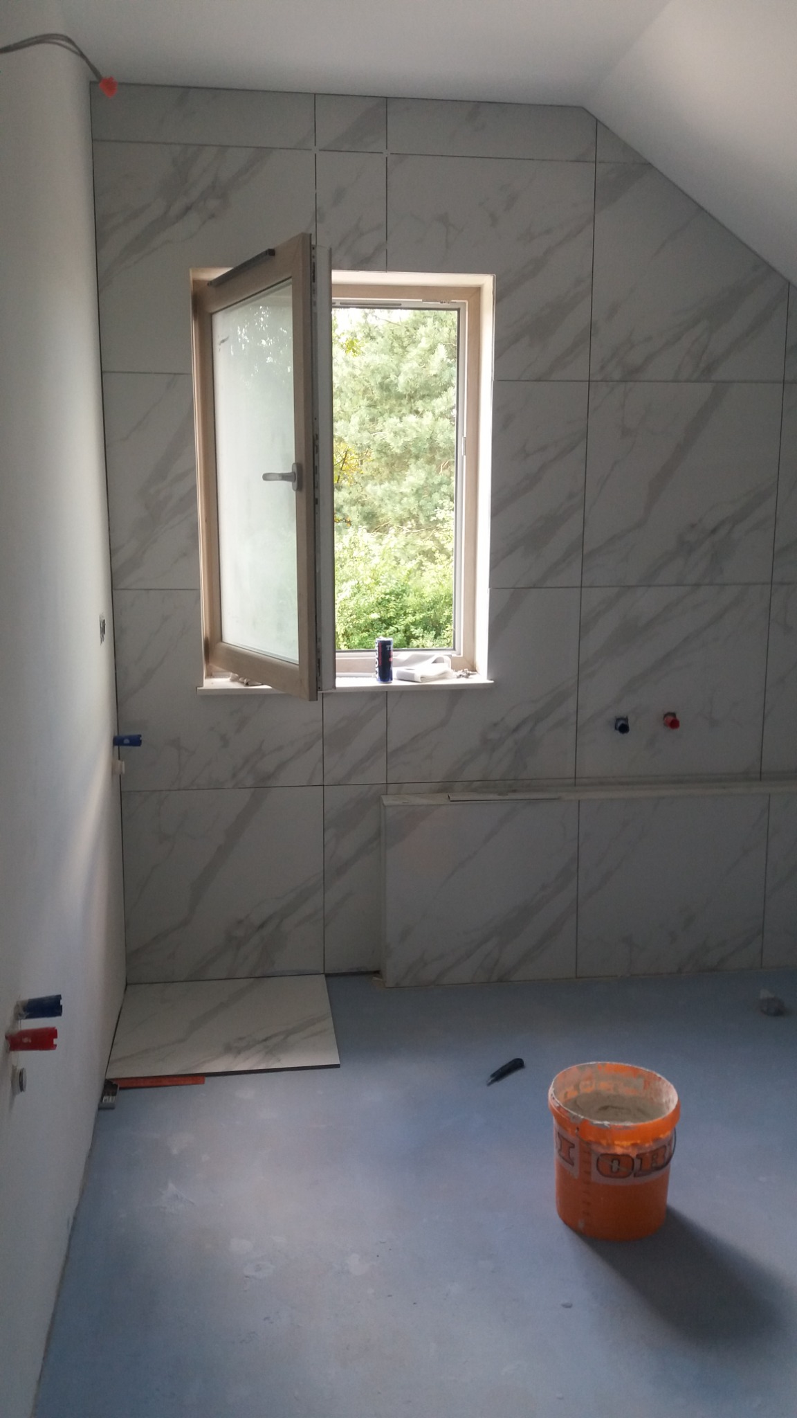 Wnętrze łazienki w trakcie prac wykończeniowych, ściany wyłożone płytkami imitującymi marmur, otwarte okno z widokiem na drzewa, na podłodze wiadro z zaprawą i pojedyncza płytka.