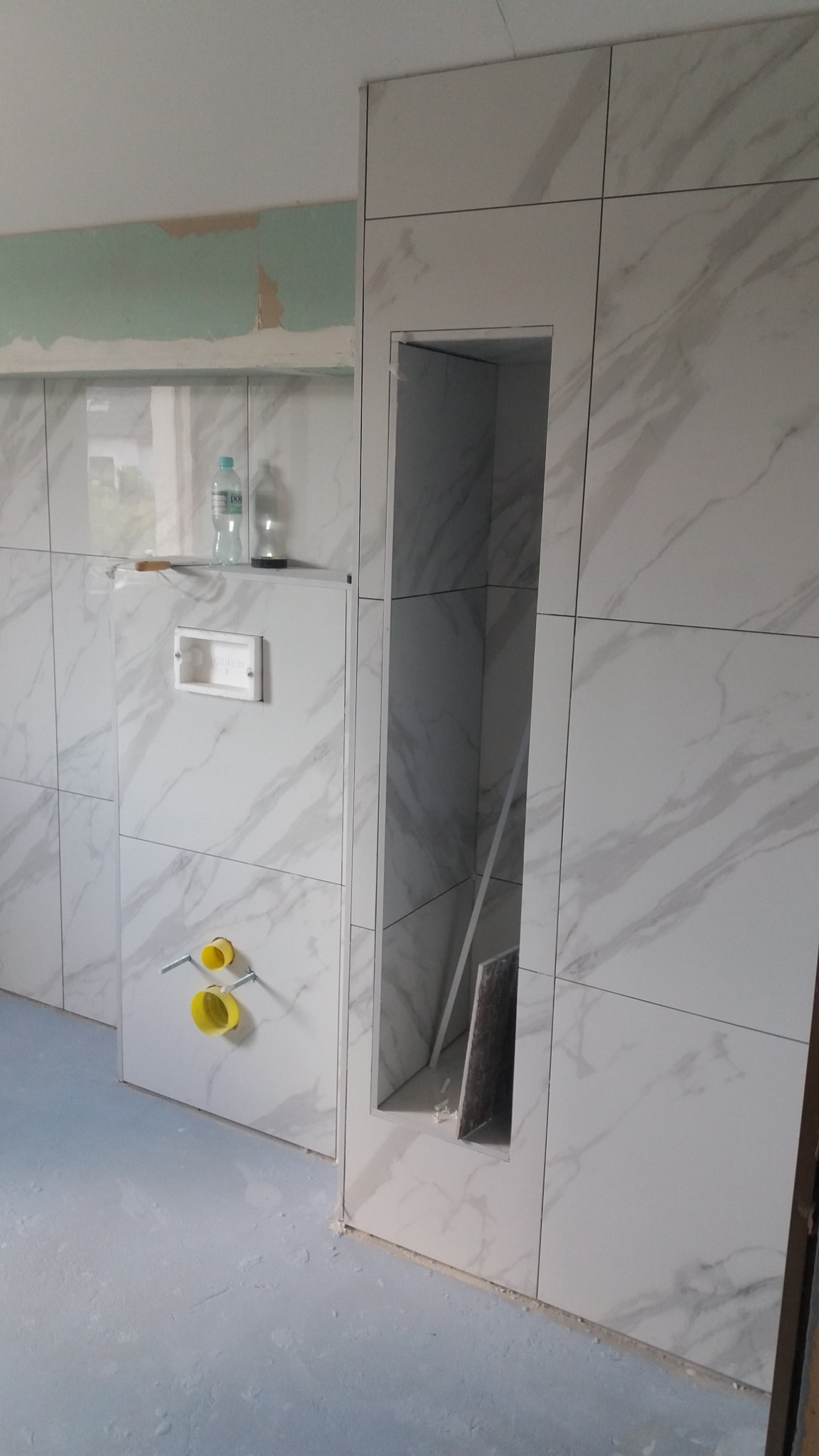 Wykończenie łazienki w trakcie remontu: białe płytki imitujące marmur na ścianach, wnęka w ścianie, żółte elementy instalacji wodnej, butelki z wodą na półce i surowy stan podłogi.