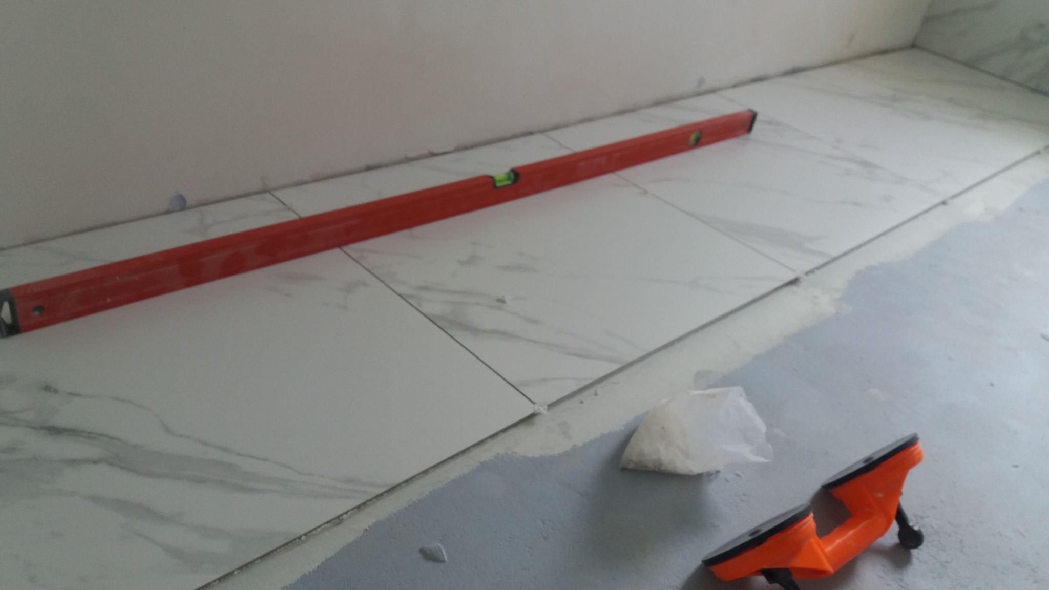 Białe płytki imitujące marmur ułożone na podłodze, poziomica w kolorze czerwonym, przyssawki do płytek i woreczek z krzyżykami dystansowymi.