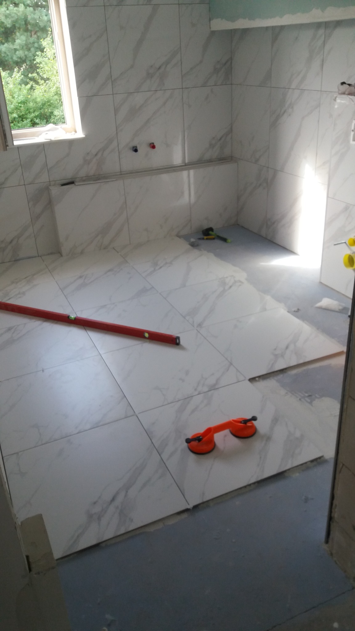 Układanie płytek podłogowych imitujących marmur w łazience, widoczna poziomica i przyssawka do przenoszenia płytek, częściowo ułożona podłoga.