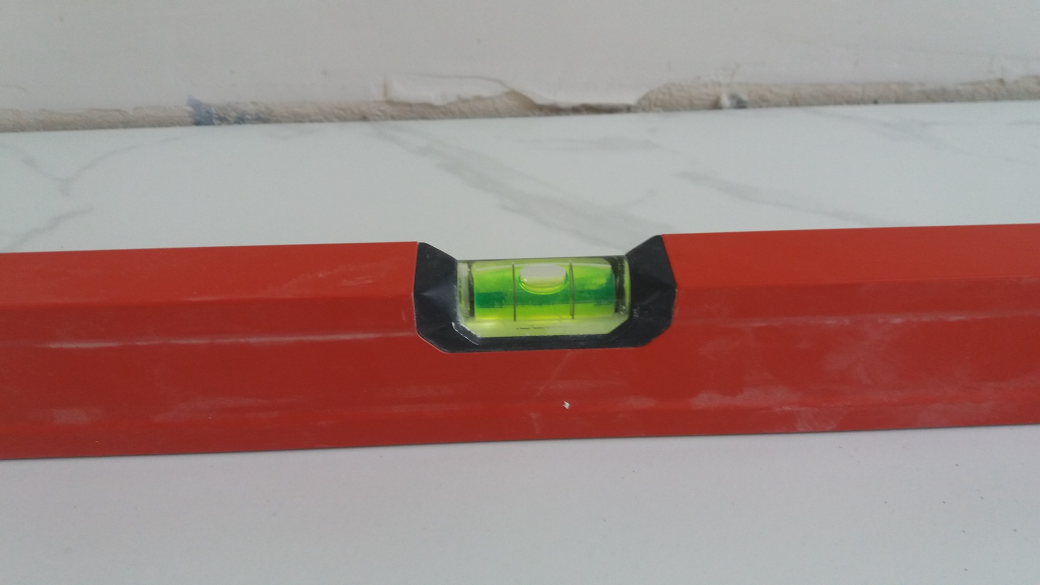 Poziomica w kolorze czerwonym, z widoczną zieloną cieczą wskazującą poziom na tle białej powierzchni i fragmentu ściany z pozostałościami po remoncie.